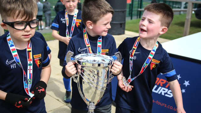 3 boys from the same team smile and chat with medals around their necks and the central boy holding a silver trophy