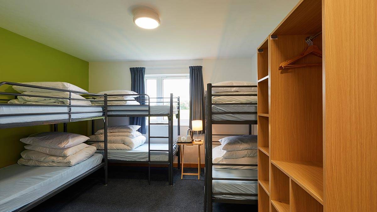 Multi-bunk bed room with three beds, a wardrobe, bed-side table and lamp and window in the background letting light in