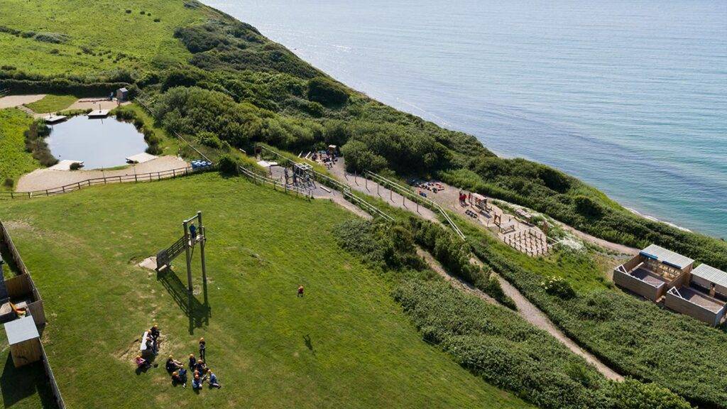 Green hills slope to the coast, with a pond and zip wire activity in the foreground and the sea and obstacle course in the backgroundthe sea