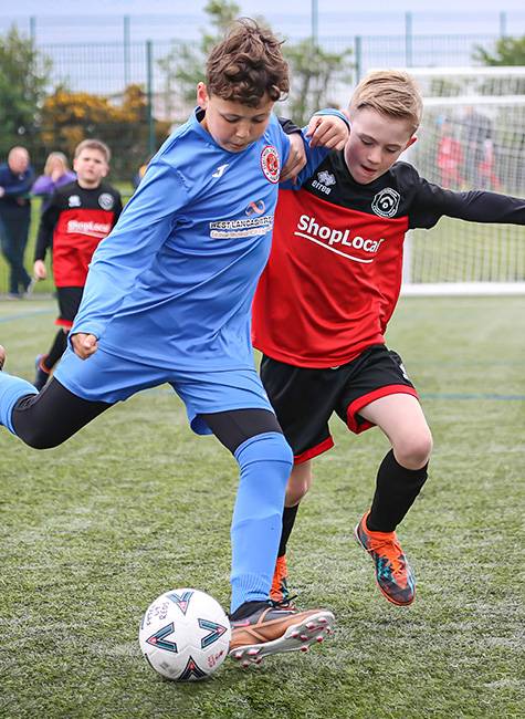 Football player in blue about to kick the ball as he's challenged by a player in red and black.