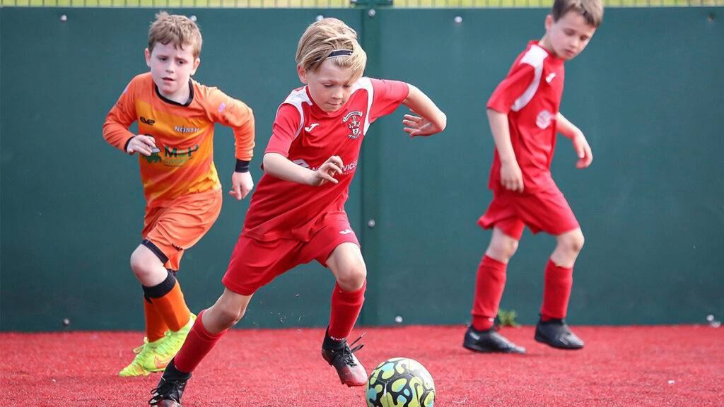 One kid dribbles with the ball on a red astro turf pitch as two other players look on