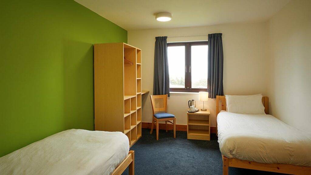 Single bed on left foreground with open cabinet storage, chairs, desk and a second single bed in the background with a window.