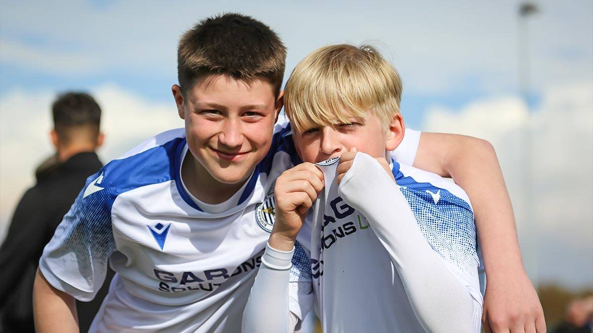 Two boys from the same team pose for the camera, with one kissing his team crest on his football jersey