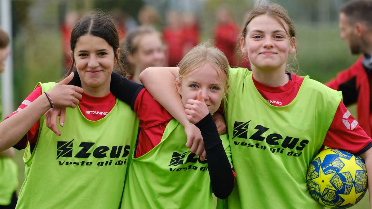 3 girls football players from a grassroots team, with arms linked around shoulders, smile, pose and put thumbs up to the camera.