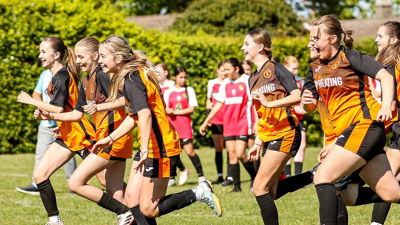 A young girls football team run up pitch celebrating after a goal